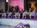 Una escena de conferencia o evento con personas sentadas en sillas blancas, algunas tomando notas. El fondo es rosa y hay plantas decorativas.