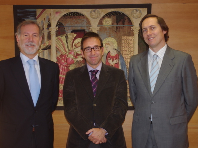 Tres hombres en trajes posan frente a un mural con motivos religiosos en un entorno formal.
