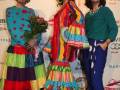 Tres mujeres posan frente a un cartel del SIMOF con vestidos de flamenco coloridos y flores.