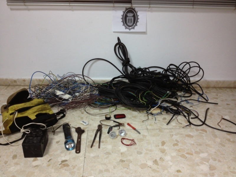Una variedad de cables y herramientas desordenadamente dispuestos en el suelo, junto con un reloj de pulsera y una batería. En la pared hay un cartel con un emblema en forma de corona.