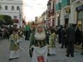 Procesión tradicional con vestimentas históricas y mantón de color verde en una calle adoquinada.