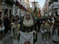 Procesión religiosa con vestimentas tradicionales y estandartes, celebrada en una calle urbana iluminada.