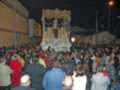Una procesión religiosa nocturna con una imagen de la Virgen en el centro, rodeada por un gran grupo de personas vestidas con ropa casual. El ambiente es festivo y el cielo está oscuro, indicando que la imagen fue tomada en la noche.