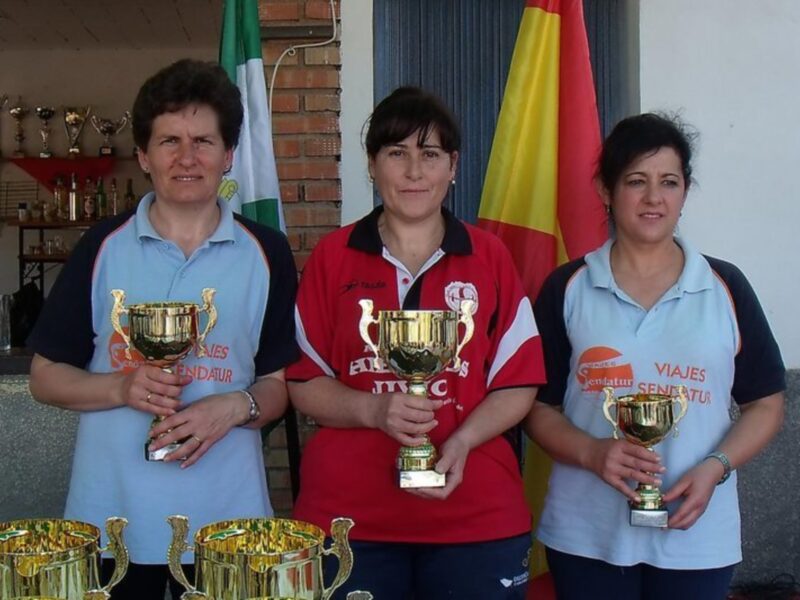 Tres personas sonrientes sostienen trofeos dorados en una celebración. Detrás de ellas, se pueden ver banderas y estantes con trofeos adicionales.