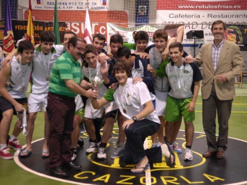 Un equipo de jóvenes jugadores de baloncesto celebrando su victoria en un campo de baloncesto, con un entrenador y varios jugadores sonriendo y posando para la foto. En el fondo, se pueden ver banderas y carteles publicitarios.