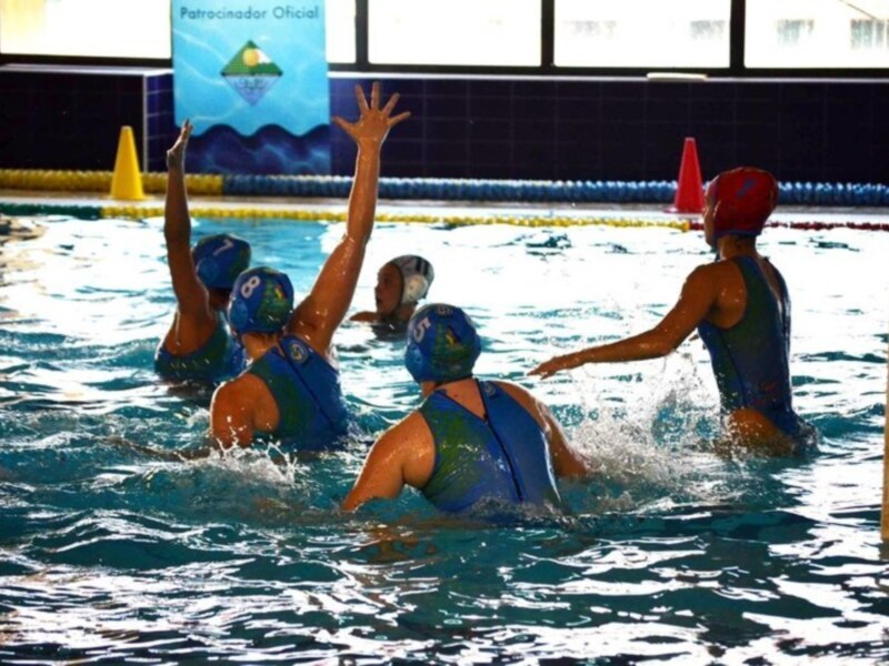 Equipo de waterpolo femenino en acción durante un partido en una piscina interior, con un patrocinador oficial en el fondo.