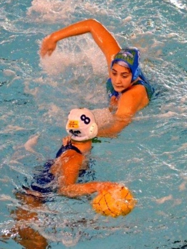 Jugadora de waterpolo en acción, con una pelota en la mano y una gorra de natación en la cabeza, nadando en un estanque de agua azul claro.