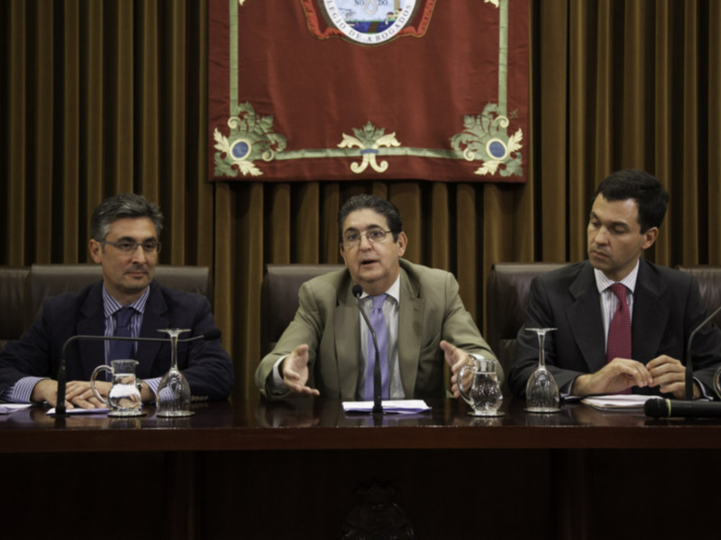Tres hombres sentados en una mesa de conferencia, con un fondo de cortinas y un escudo heráldico en la pared.