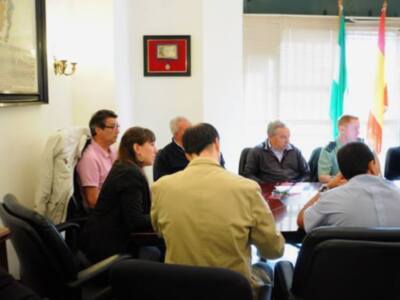 Reunión de trabajo en una sala de conferencias con personas sentadas alrededor de una mesa. La pared está decorada con una bandera y un marco de fotos, mientras que la iluminación es natural a través de las ventanas.