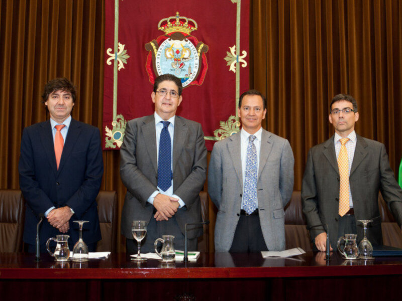Los hombres están posando en una mesa frente a un fondo con un escudo y una corona, probablemente en un evento oficial o de negocios.