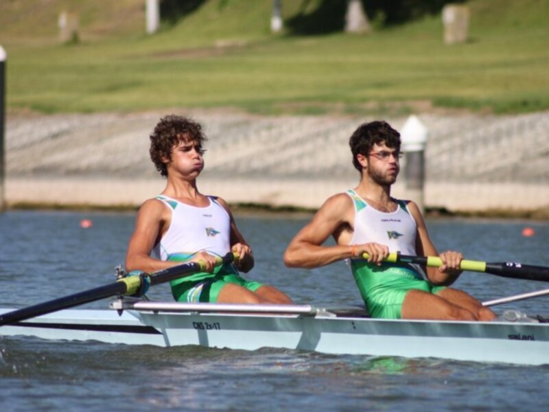 Dos personas remando en un bote de remo en un lago, con un fondo de césped y muelles en el horizonte.