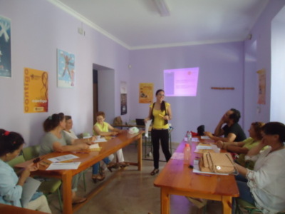 Una sala de conferencias con personas sentadas en mesas, una mujer en el centro presentando algo en un proyector. La sala tiene carteles y una caja de herramientas colgada en la pared.