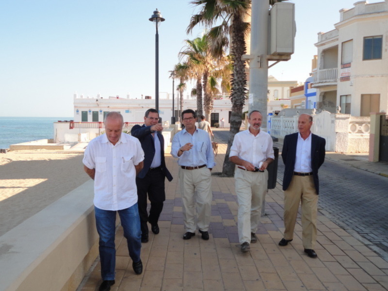Grupo de personas caminando en una calle con palmeras y edificios al fondo, en un entorno urbano con una vista de la playa en el fondo.