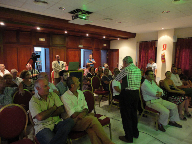 Auditorio con personas sentadas en sillas rojas, uno de ellos está en pie hablando. El ambiente parece ser una conferencia o presentación formal.
