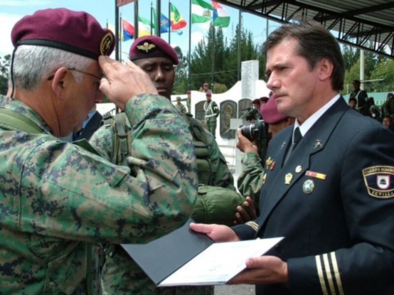 Un oficial militar entrega un documento a un soldado en una ceremonia militar. En el fondo, se pueden ver banderas y una estructura de metal.