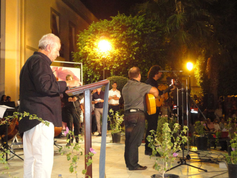Un hombre en un traje negro habla frente a una pizarra blanca con imágenes, mientras otro hombre toca la guitarra eléctrica en un escenario iluminado por faroles. La escena se desarrolla al aire libre, con plantas y árboles en el fondo.