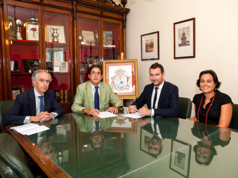 Un grupo de personas sentadas en una oficina con un escritorio grande y varios estantes llenos de objetos. La pared detrás está decorada con cuadros y una vitrina con trofeos.
