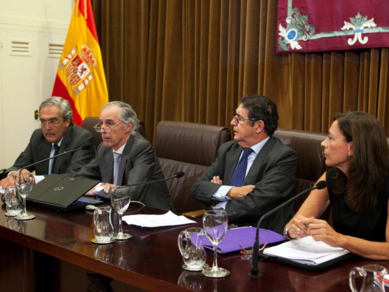 Sesión de trabajo en una sala de conferencias con tres personas sentadas alrededor de una mesa, entre ellos un hombre y dos mujeres. La bandera española se encuentra a la izquierda del grupo, y una bandera roja con un emblema en el fondo. La mesa tiene portátiles y documentos, indicando una reunión formal o de negocios.