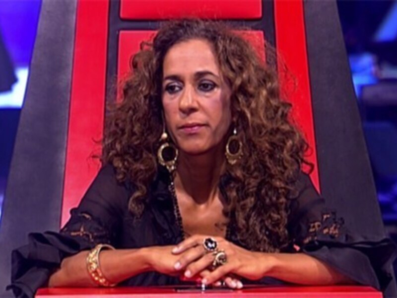 A una mujer sentada en un asiento rojo, posiblemente en un programa de televisión. La escena parece ser de una competencia o concurso, dado el entorno y la vestimenta formal.