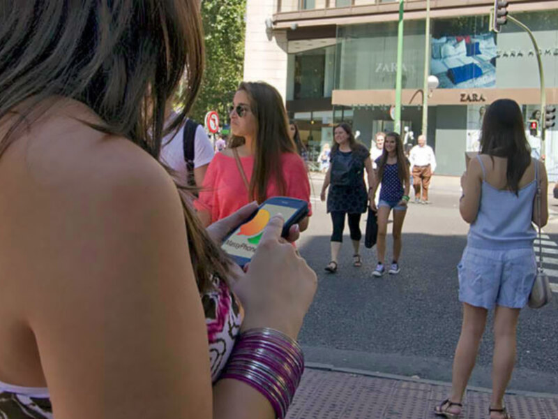 Una mujer con un vestido rosa y una camiseta blanca, usando un teléfono móvil, camina por una calle con otros peatones. La imagen muestra una calle con edificios comerciales y un semáforo en el fondo.