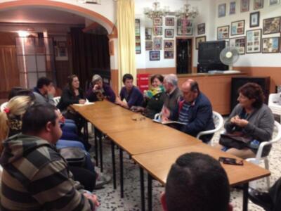 Grupo de personas reunidas en una sala con mesas y sillas, discutiendo. En la pared hay cuadros y una pantalla de televisión en el fondo. La iluminación es natural, proveniente de ventanas no visibles.