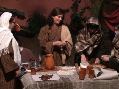Una imagen de una escena navideña con personas vestidas con abrigos y mantas, sentadas alrededor de una mesa cubierta con un mantel. La mesa tiene varios platos, incluyendo galletas y posiblemente dulces navideños. La atmósfera parece ser festiva, probablemente una celebración o reunión familiar en torno a la Navidad.