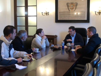 Un grupo de personas reunidas en una sala de conferencias. En la mesa hay tazas de café y documentos. La pared está decorada con un cuadro que parece ser una bandera o escudo histórico.
