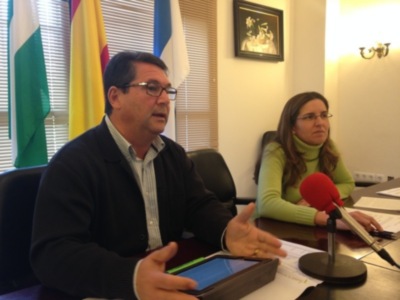 En una sala de conferencias, un hombre habla en un micrófono rojo mientras una mujer asiste a la reunión. Detrás de ellos, se ven banderas y un cuadro en la pared.