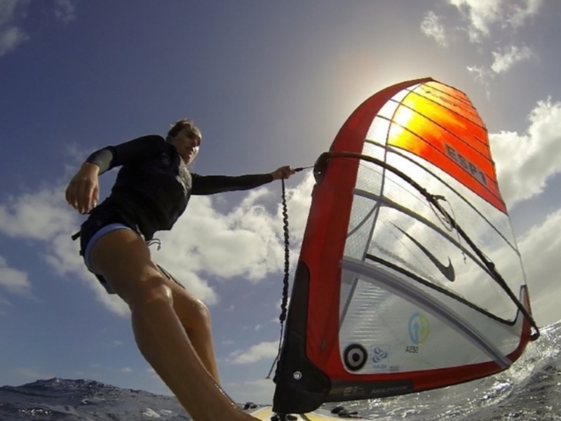 Una persona windsurfando con una vela roja y naranja en el mar, bajo un cielo azul con algunas nubes. La persona está sujetando la vela con una cuerda.