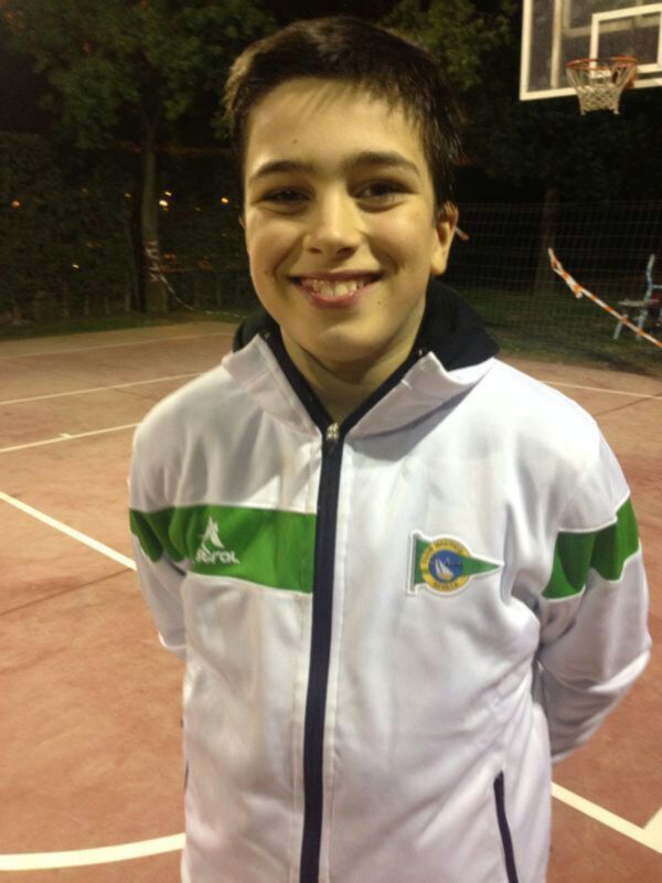 Un niño sonríe en una cancha de baloncesto, vestido con una chaqueta blanca con detalles verdes y azules, con un escudo en la parte delantera que parece representar a Brasil.