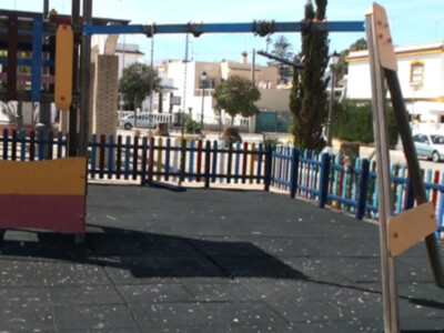 Un parque infantil con columpios y un muro de madera en el centro, rodeado por un pequeño muro de madera de colores y un camino de asfalto. El cielo está despejado y hay edificios en el fondo.