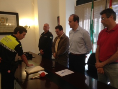 Un policía en uniforme amarillo está frente a una mesa, con un portátil abierto y un micrófono en el suelo. Algunos hombres observan la escena, uno de ellos con una chaqueta marrón y gafas. La imagen muestra un ambiente formal, posiblemente una conferencia o reunión de trabajo.