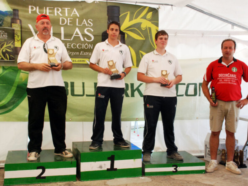 Podio de una competición con tres participantes en el centro y un juez a la derecha, bajo un cartel de 'Puerta de las Villas'.