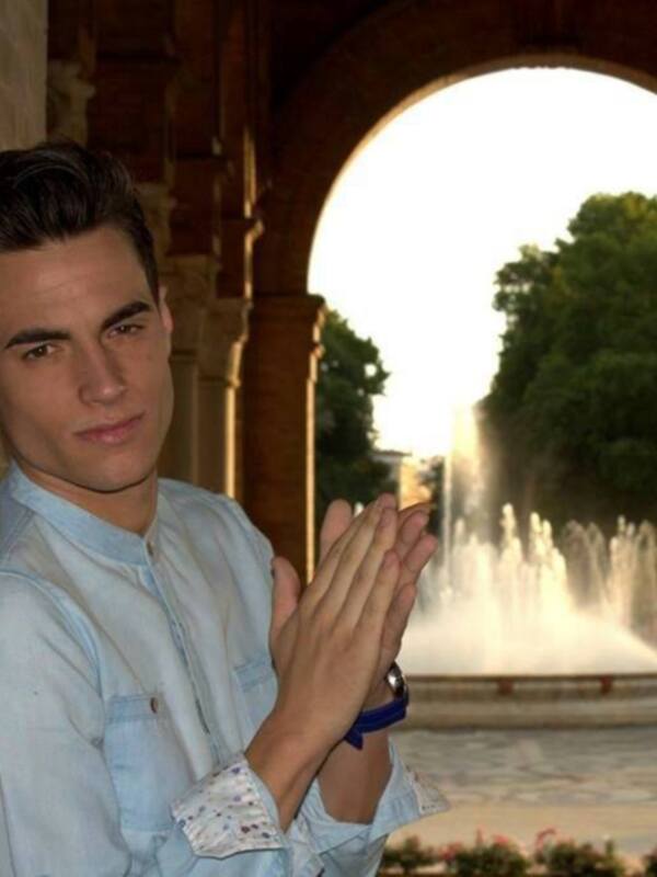 Un hombre joven con una camisa azul claro posa con las manos juntas frente a un arco y una fuente en el fondo.