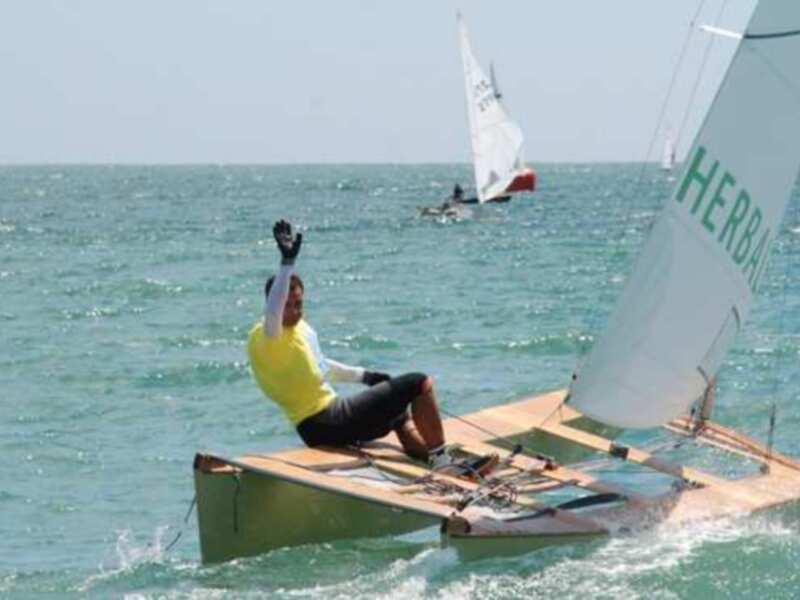 Un regatista celebra su victoria en una regata de vela, con otro barco a la distancia bajo un cielo azul claro.