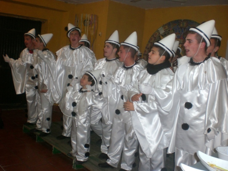 Un grupo de niños vestidos con trajes blancos y negros, posiblemente para una celebración o fiesta. El ambiente parece ser interior y decorado con colores amarillos y marrones, sugiriendo una festividad o evento familiar.