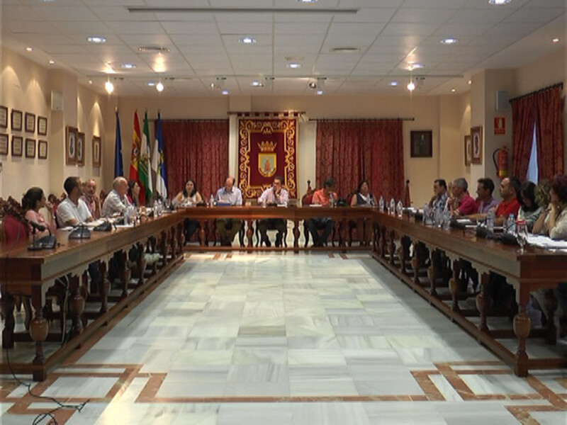 Sala de reuniones con una mesa larga y varios asistentes sentados. La pared tiene varias banderas y un escudo heráldico. El suelo es de mármol con patrones geométricos.