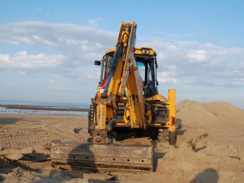 Una excavadora amarilla está trabajando en una playa, con arena y dunas alrededor. El cielo es azul con algunas nubes.