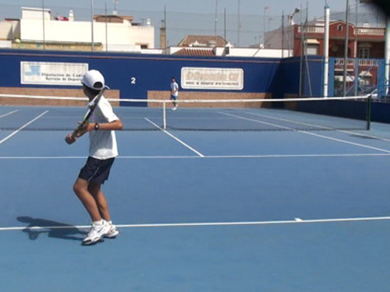 Un joven con un casco blanco y una camiseta blanca está listo para jugar tenis en una cancha azul. La imagen muestra la parte superior de su cuerpo, incluyendo sus piernas y manos. El fondo muestra una línea de cancha azul con líneas blancas, un poste de red y edificios en el fondo.