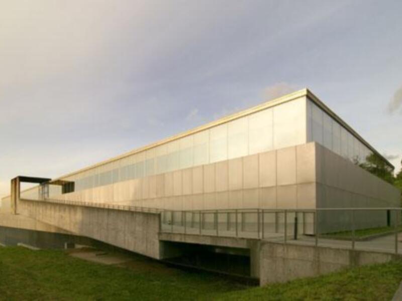 Edificio moderno con fachada de vidrio y madera, ubicado en un entorno verde con árboles y césped.