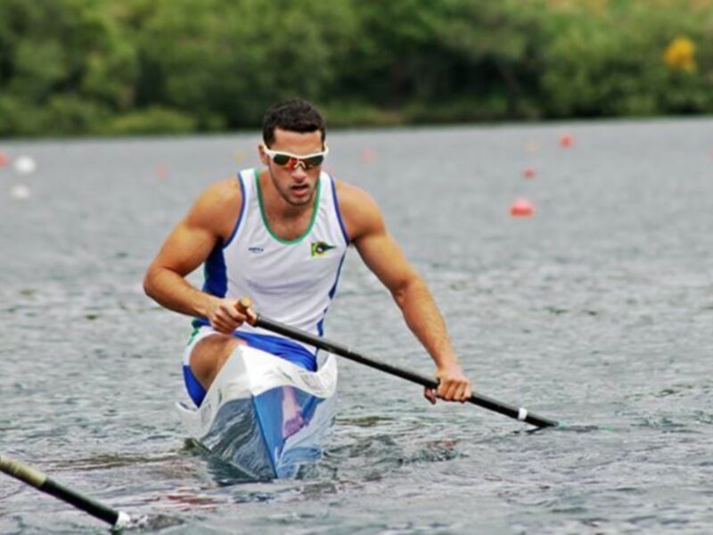 Atleta brasileño compitiendo en una regata de piragüismo en un lago rodeado de árboles.