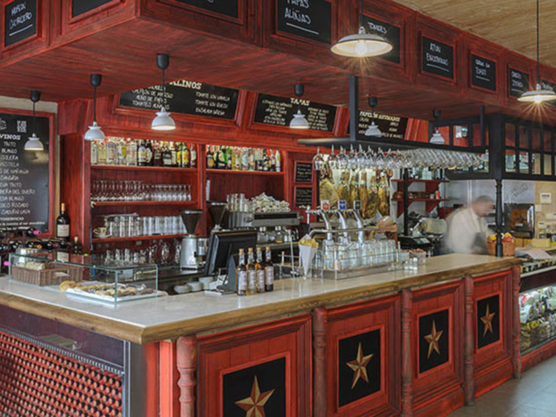 Bar con mostrador rojo y estrellas doradas, mesas de madera y botellas en estanterías.