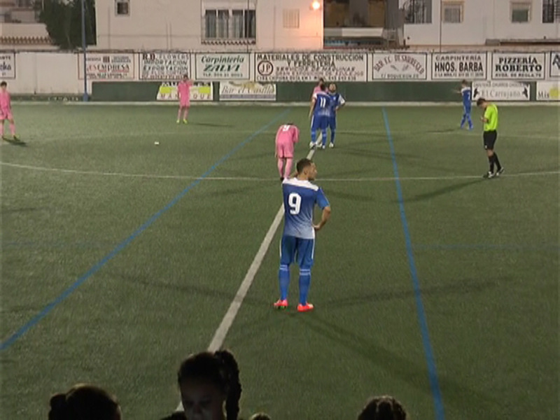 Un jugador de fútbol en azul y rojo se prepara para un tiro libre, con el campo de juego al fondo y publicidad en las paredes.