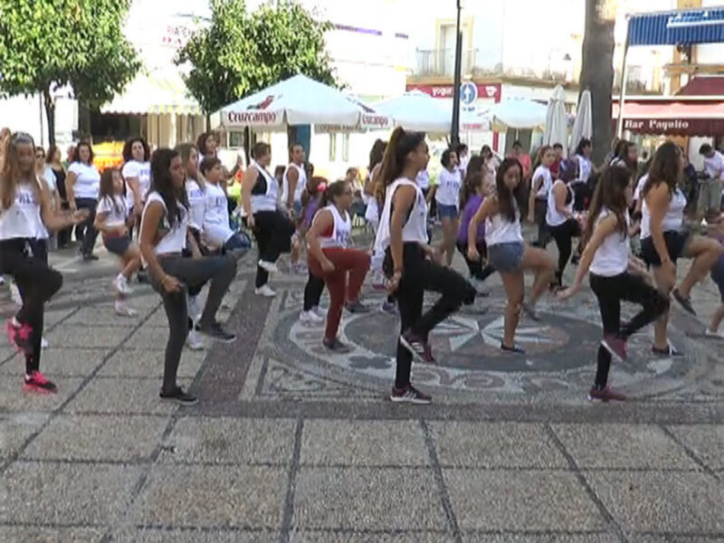 Una multitud de personas realiza una coreografía en un parque público. La escena está rodeada por árboles y edificios comerciales en el fondo.