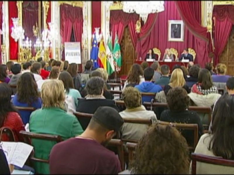 Una audiencia en un salón con cortinas rojas y un escenario con personas sentadas en sillas. En el fondo, se puede ver una bandera española y una imagen de un hombre en el escenario. Los asistentes están concentrados en la presentación.