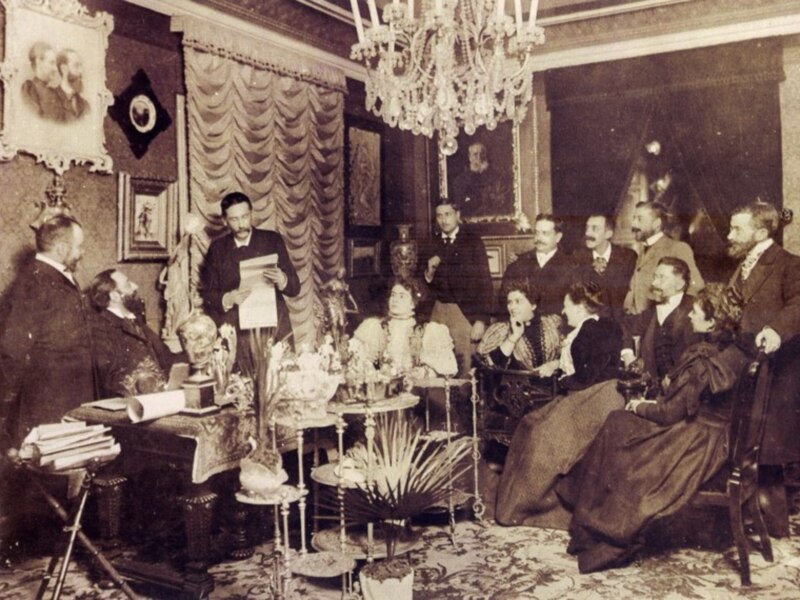 Una reunión formal en un salón elegante con cortinas y un candelabro. Las personas están vestidas con trajes de época, lo que sugiere una escena histórica o ceremonial. La decoración y el estilo de vestimenta indican un ambiente formal, posiblemente una reunión importante o una celebración.