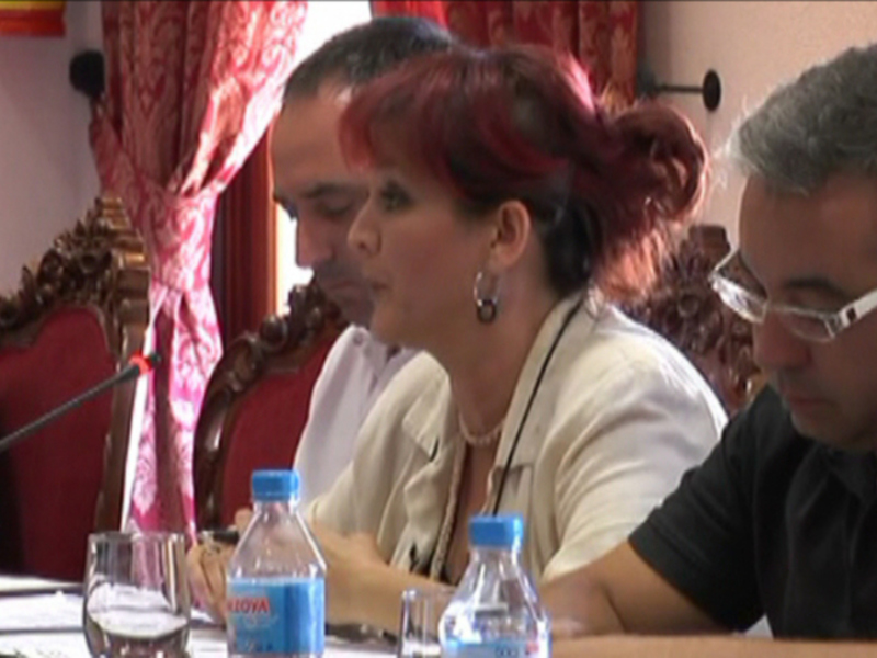 Una imagen de una reunión formal con personas sentadas en mesas, sosteniendo documentos y bebiendo agua de botellas azules. La sala tiene cortinas rojas y una silla con un ornamento dorado en el fondo.