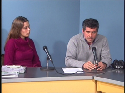 Dos personas sentadas en una mesa con documentos y micrófonos, probablemente en un evento formal o conferencia.