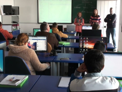 Clase de informática en un aula moderna con estudiantes sentados frente a sus computadoras y una profesora explicando en el frente.