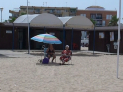 Dos personas sentadas bajo un paraguas multicolor en la playa, con casetas y edificios al fondo.